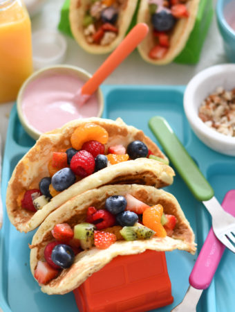 A white table background with rainbow pancake tacos, oj, and silverware.